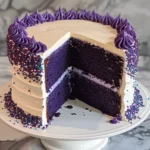 A sliced purple velvet cake with cream cheese frosting, decorated with purple and multicolored sprinkles, displayed on a white cake stand.