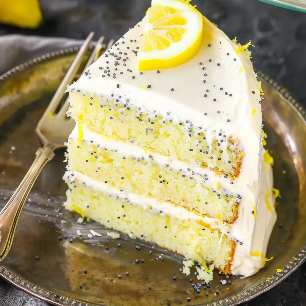 A slice of lemon poppy seed cake with creamy white frosting and lemon garnish on a silver plate.